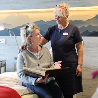 Eine Pflegekraft steht neben einer sitzenden Person im Gästezimmer des Oberland Hospiz, legt ihr beruhigend die Hand auf die Schulter.